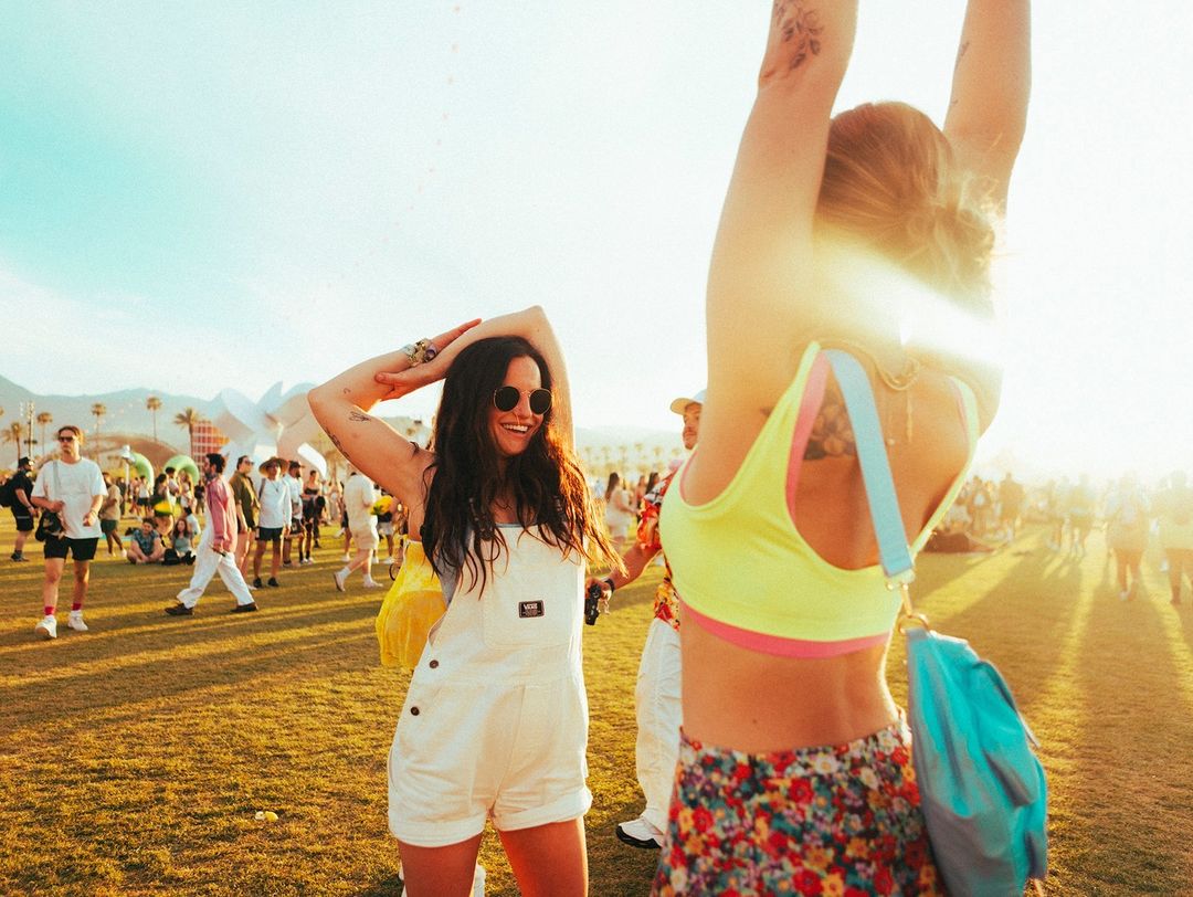 Two adult women smile outdoors on a grassy field at a music festival on a sunny day, wearing summery casual outfits and sunglasses.