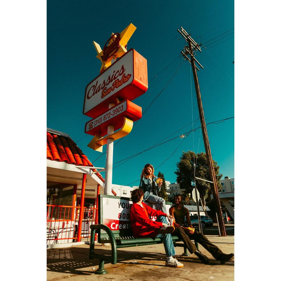 Three young adults relax on a park bench under a vintage 'Classics San Pedro' sign on a sunny day, enjoying food and drinks together.