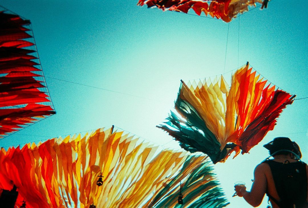 Low-angle shot of colorful hanging decorations under bright blue sky, with a person visible in the lower right corner holding a glass.