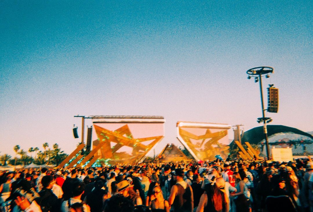 Large crowd of people gathered at an outdoor music festival in front of a stage with large screens under a clear, sunny sky.
