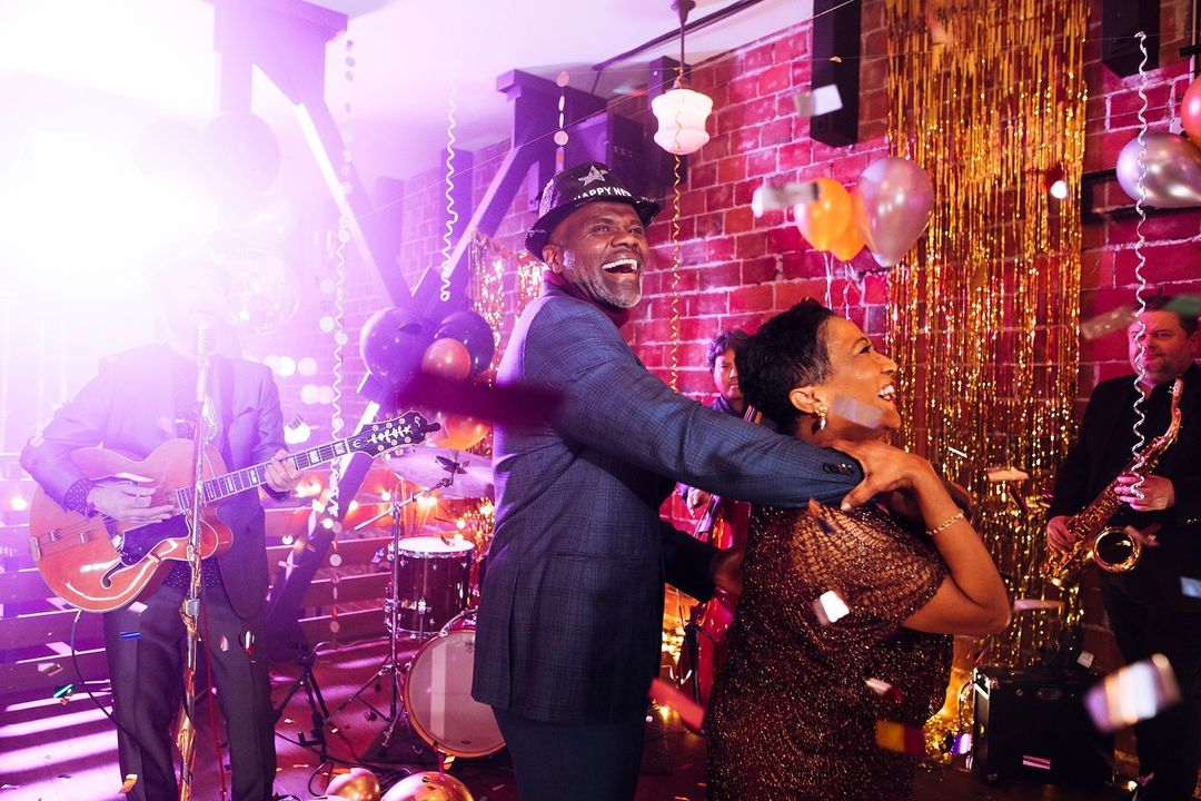 Enthusiastic couple dancing with live band in festive party setting, confetti falling, lit by vibrant lighting. New Year's Eve celebration.