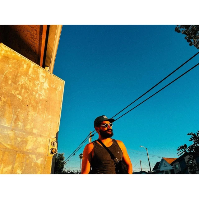 Adult male with beard and sunglasses poses in gray tank top in outdoor urban setting under clear blue sky near concrete structure.