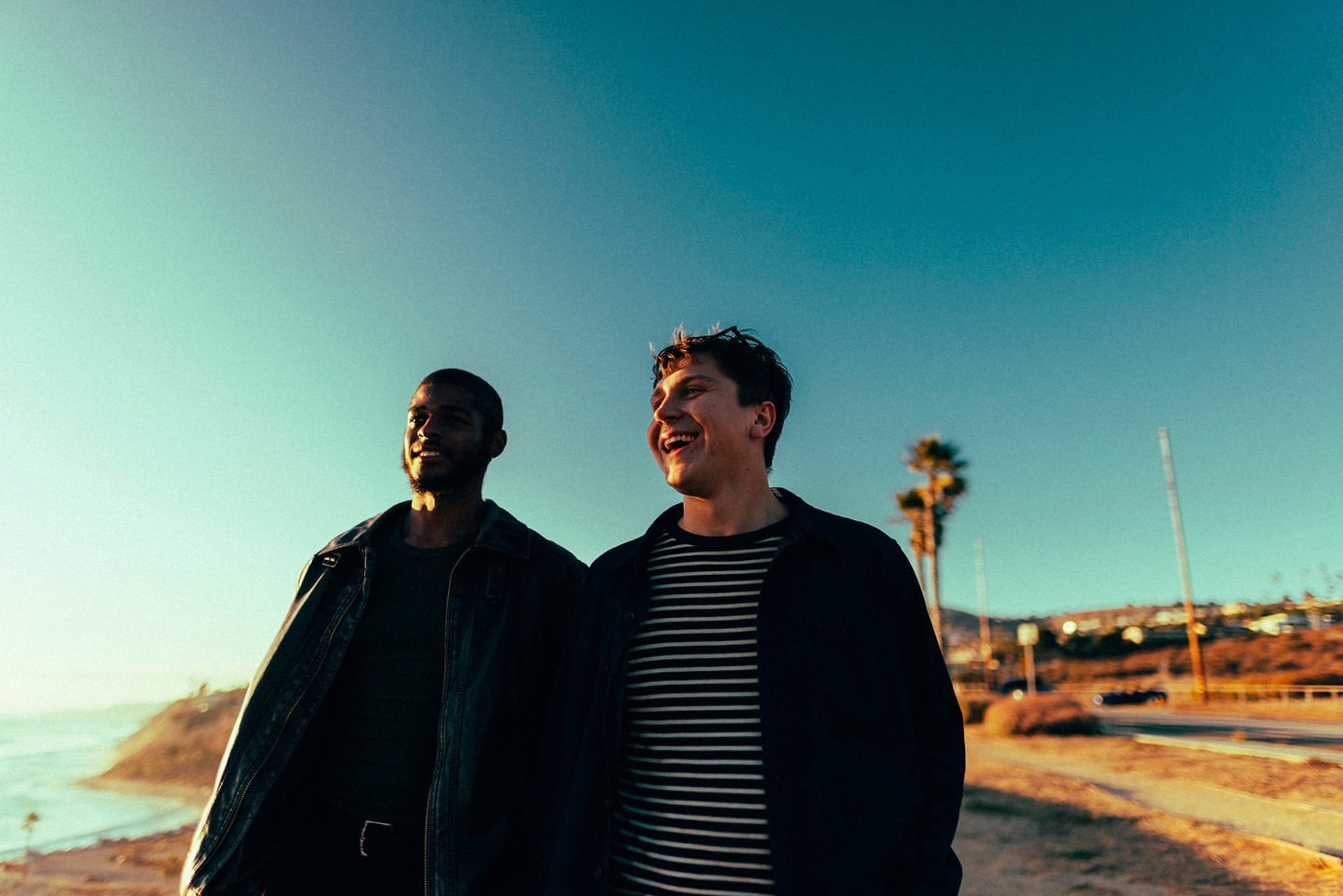 Two young men walk along a beachside path under a blue sky. One man is wearing a striped shirt, and the other a dark jacket.