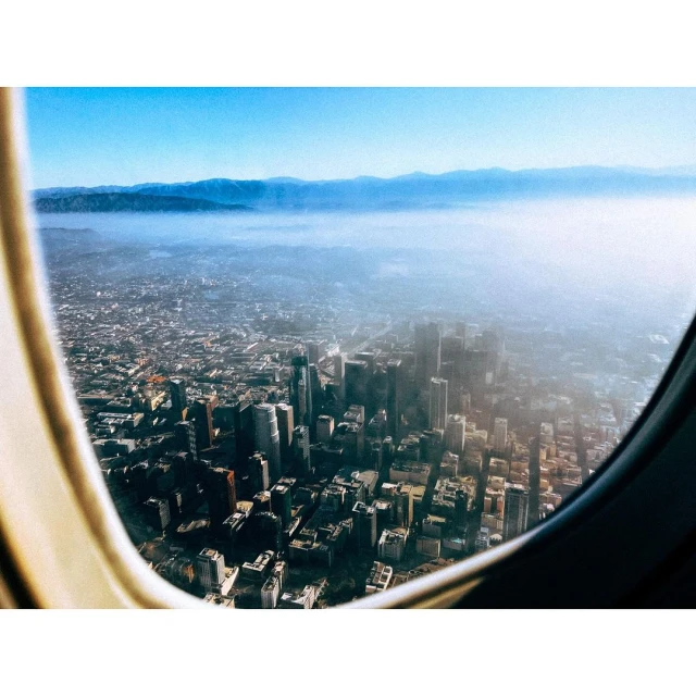 Airplane window view of a city skyline under a blue sky with clouds, showcasing urban landscape and travel photography.