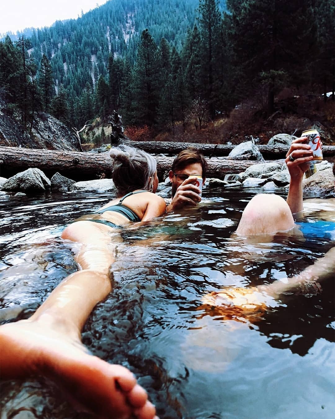 Three people, two adult females and one adult male, enjoy beers while sitting in a shallow river with a forest background.