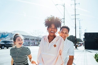 Three young adults are walking on a sunny street, smiling and laughing with building and overhead power lines in the background.