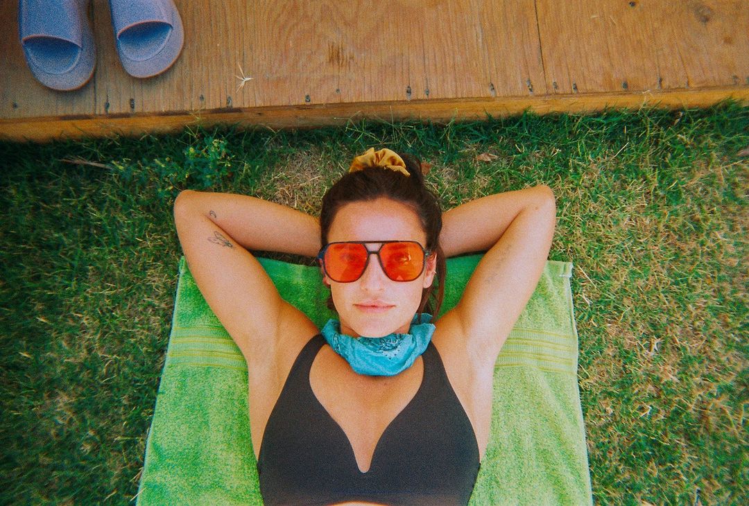 Woman lies on a green towel in the grass wearing sunglasses and a bandana, with a wooden deck and sandals in the background.
