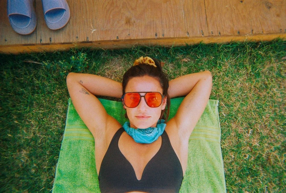 Woman lies on a green towel in the grass wearing sunglasses and a bandana, with a wooden deck and sandals in the background.