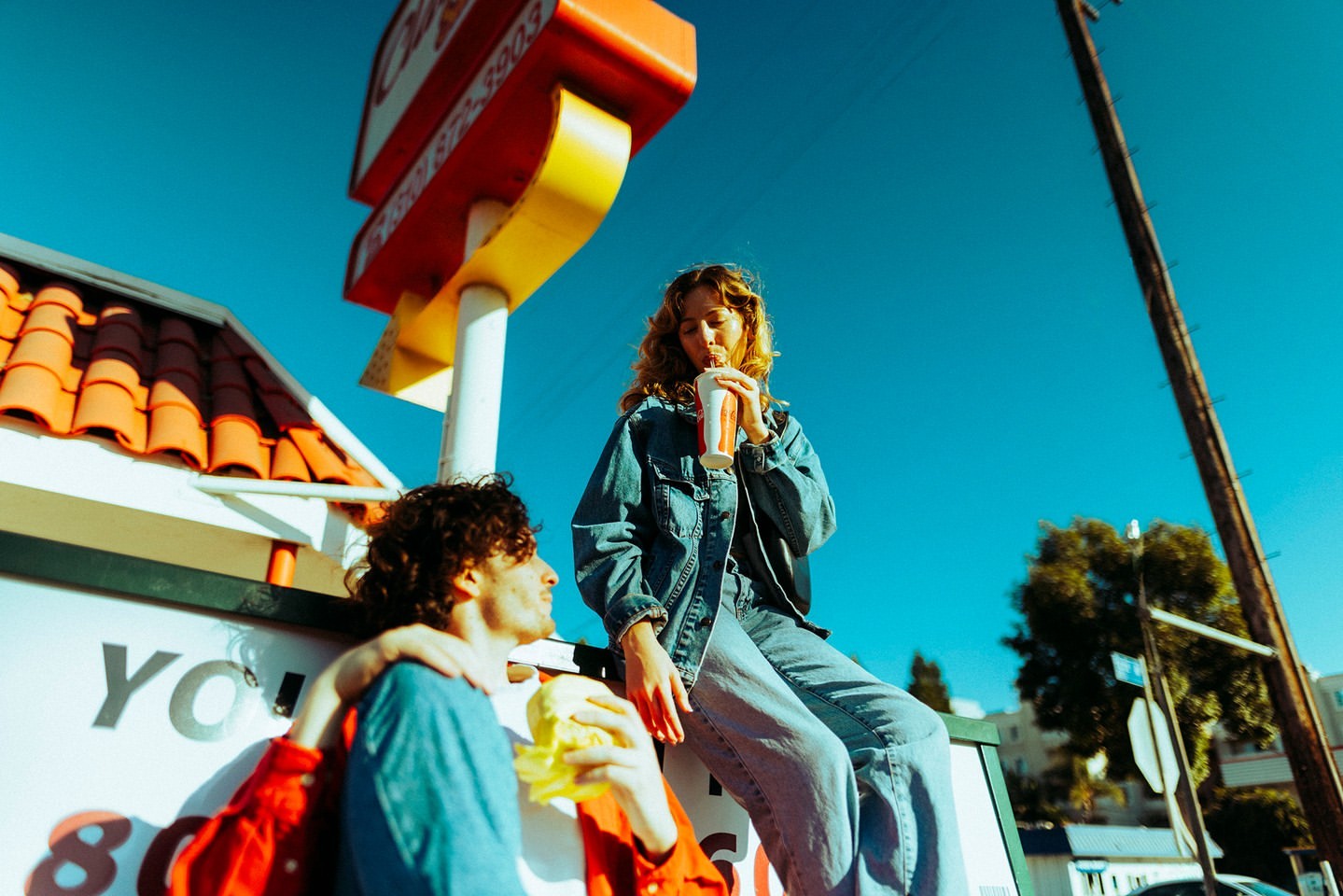 Two people outside a restaurant with a vintage sign. One is drinking from a cup, the other eating, under a clear blue sky.