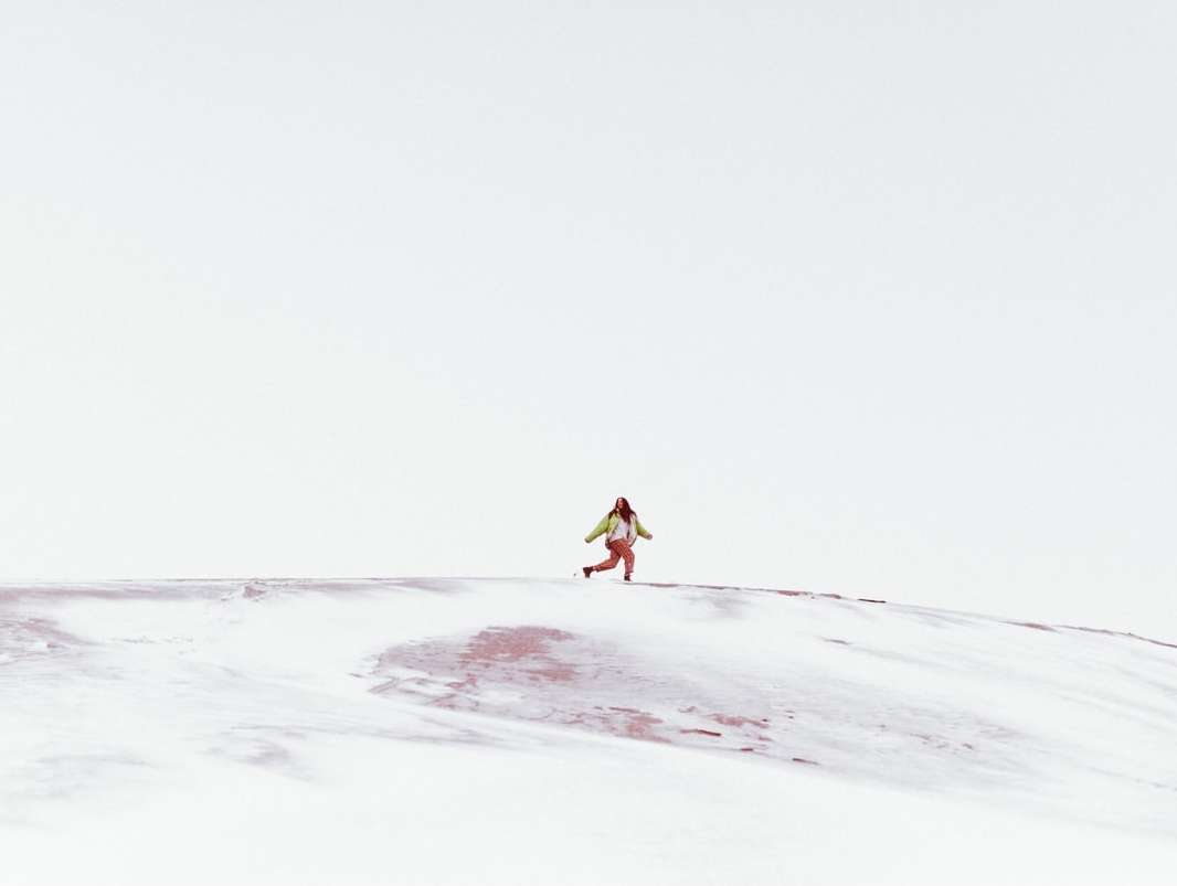 A lone woman runs across a snowy hill in a bright, minimalist landscape, wearing an olive jacket and pink pants under a clear sky.