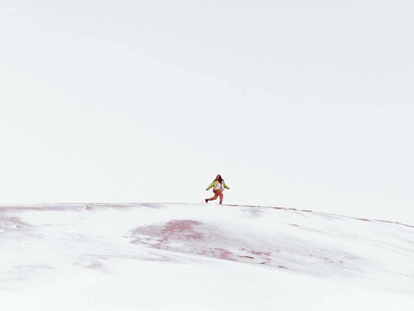 A lone woman runs across a snowy hill in a bright, minimalist landscape, wearing an olive jacket and pink pants under a clear sky.