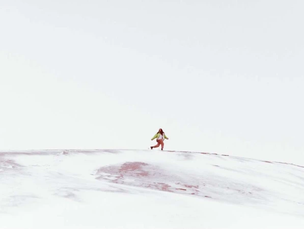 A lone woman runs across a snowy hill in a bright, minimalist landscape, wearing an olive jacket and pink pants under a clear sky.