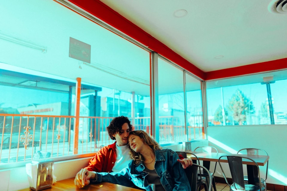 Young couple embraces in a sunlit diner, holding hands at a wooden table, showcasing intimacy and connection against a backdrop of city life.