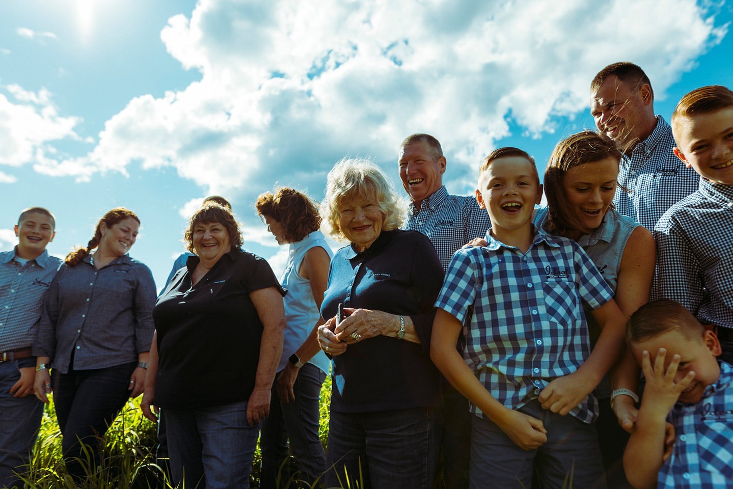 Low angle group portrait of a multi-generational family standing outdoors in a grassy field beneath a blue sky.