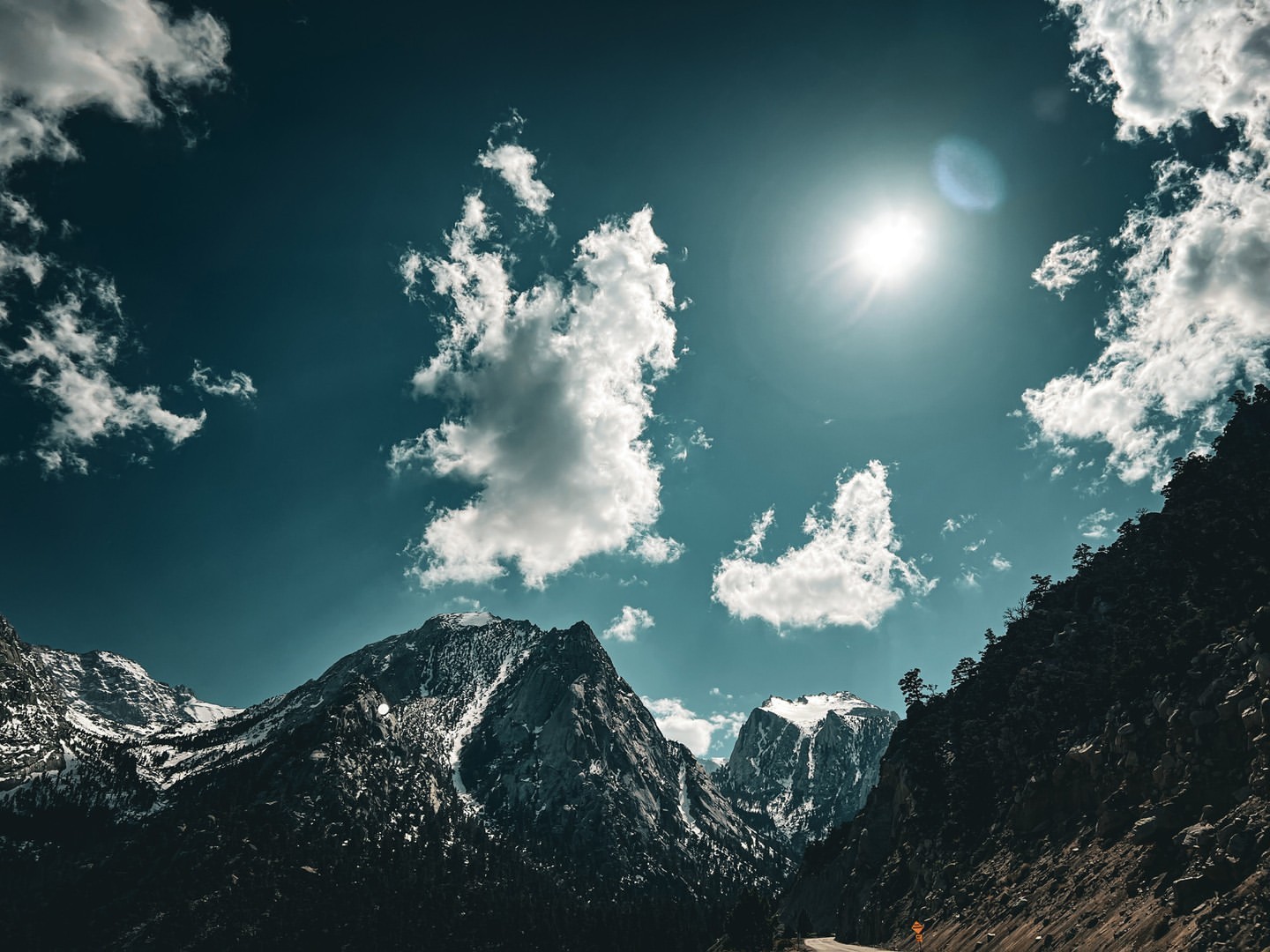 Scenic mountain landscape featuring snow-capped peaks under a blue sky with fluffy white clouds and the bright sun shining above.
