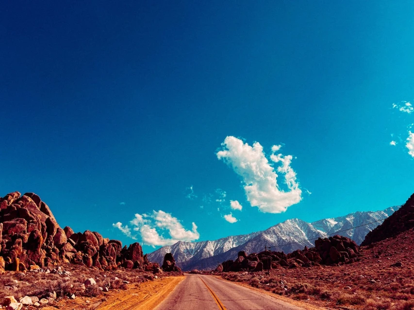 Scenic road stretches through a desert landscape with mountains in the background under a bright blue sky with scattered clouds.