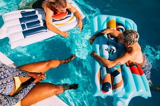 Aerial view of three people floating on inflatable rafts in a swimming pool with clear, rippling water on a sunny day.