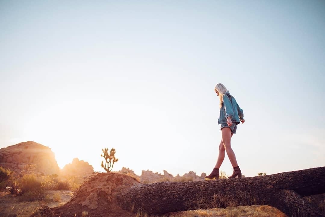 Woman with blonde hair walks across a fallen tree in a desert landscape at sunset, wearing denim jacket, shorts, and brown boots.