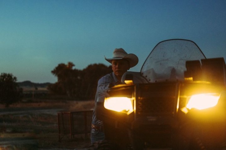 Man in cowboy hat stands next to a lit four-wheeler in a rural setting at dusk, illuminated by the vehicle's bright headlights.