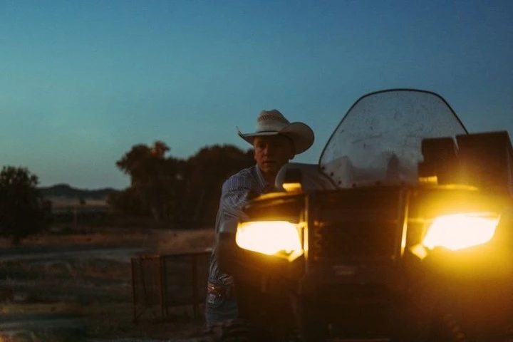 Man in cowboy hat stands next to a lit four-wheeler in a rural setting at dusk, illuminated by the vehicle's bright headlights.