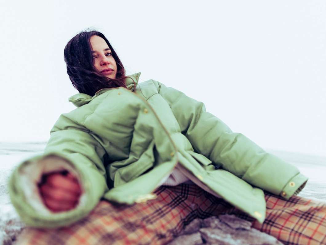 Woman in a puffy sage green coat reclines on rocks, wearing plaid pants against a bright, overcast backdrop for a fashion editorial.