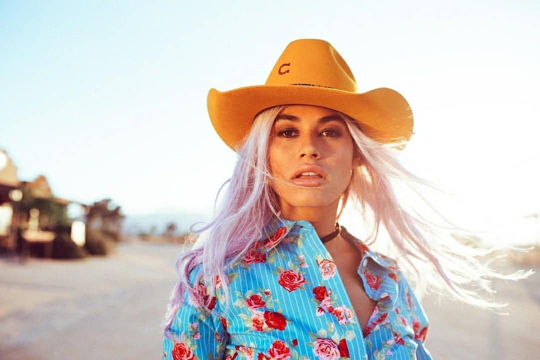 Young woman in a cowboy hat and a floral shirt in a desert landscape setting with vintage buildings in the background.