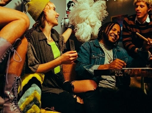 Candid shot of a group of young adults laughing together indoors, featuring diverse hairstyles, clothing and an assortment of accessories.
