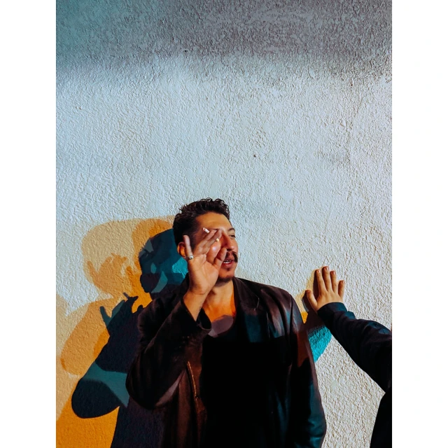 Portrait of a man with a cigarette, hand up to his face, wearing a jacket, standing in front of a white textured wall with shadows.