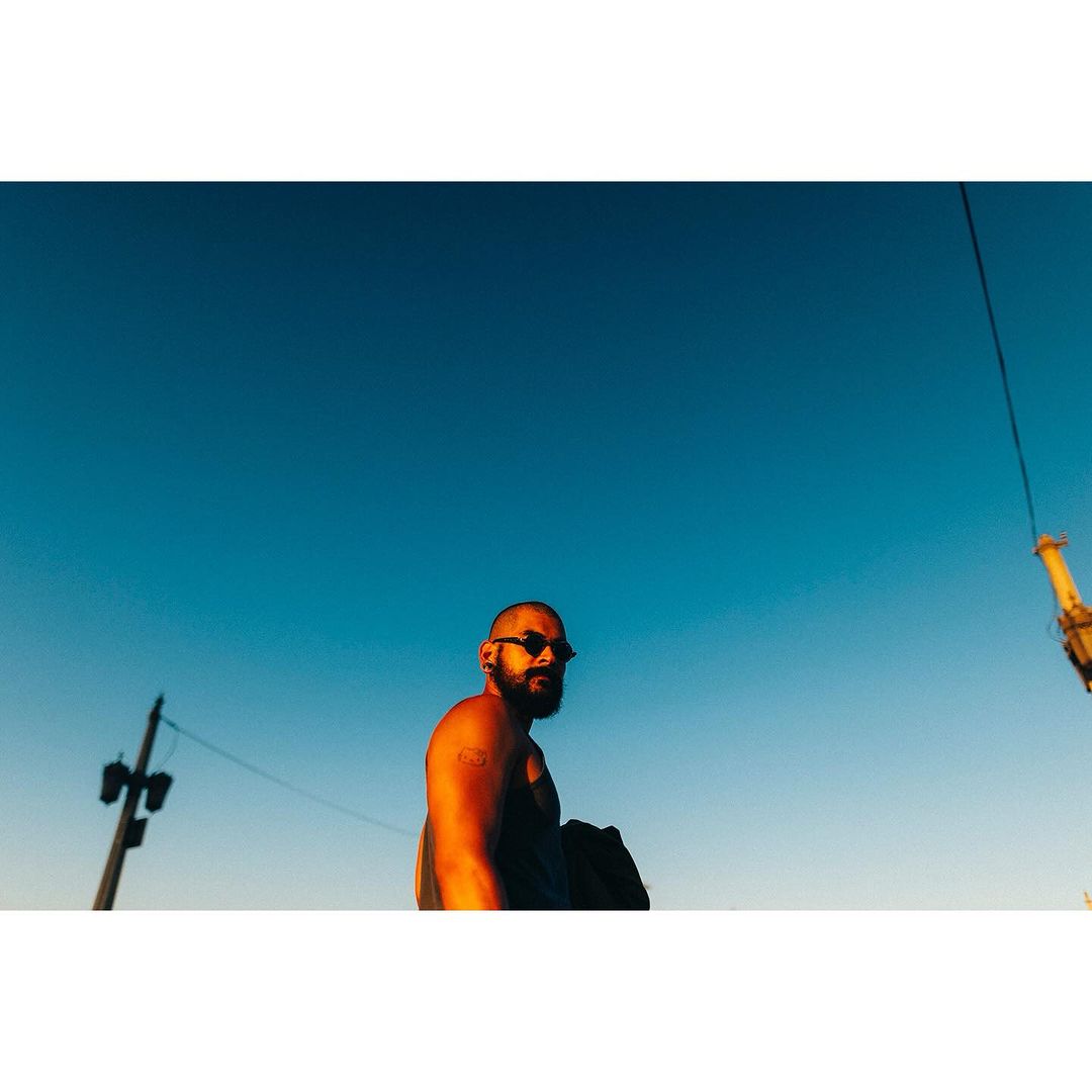 Man with sunglasses and beard standing outdoors under a clear blue sky, captured with a low angle and golden light.