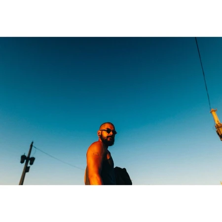 Man with sunglasses and beard standing outdoors under a clear blue sky, captured with a low angle and golden light.