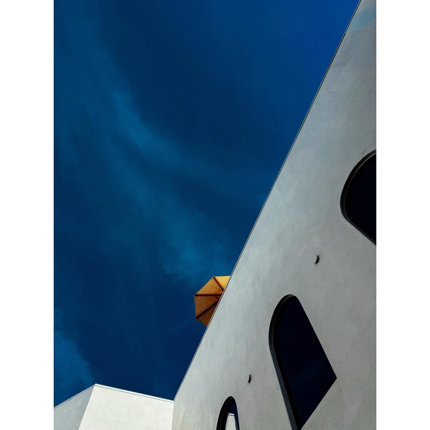 Architectural shot of a white building with arched windows and a tan umbrella against a deep blue sky with light wispy clouds.