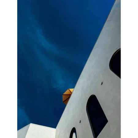 Architectural shot of a white building with arched windows and a tan umbrella against a deep blue sky with light wispy clouds.