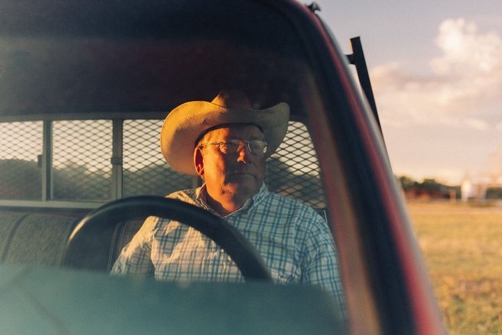 Man wearing a cowboy hat in the driver's seat of a red pickup truck with a rural field and cloudy sky visible in the background.
