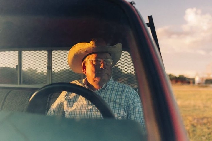 Man wearing a cowboy hat in the driver's seat of a red pickup truck with a rural field and cloudy sky visible in the background.