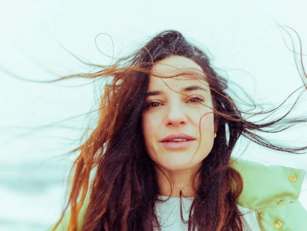 Close-up portrait of a woman with long dark windswept hair wearing a light green jacket and white top against a pale sky.