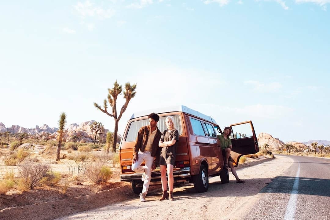 Three people and a vintage brown van are parked on a desert roadside with a Joshua Tree-filled landscape under a clear blue sky.