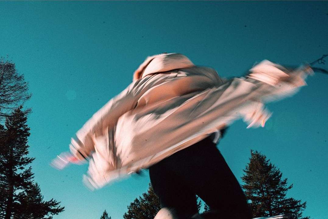 Person wearing a beige jacket jumps high in the air against a blue sky backdrop, framed by evergreen trees, showcasing motion blur.