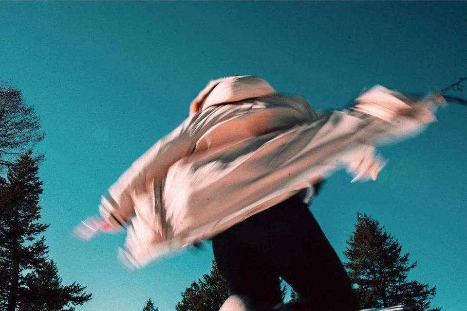 Person wearing a beige jacket jumps high in the air against a blue sky backdrop, framed by evergreen trees, showcasing motion blur.