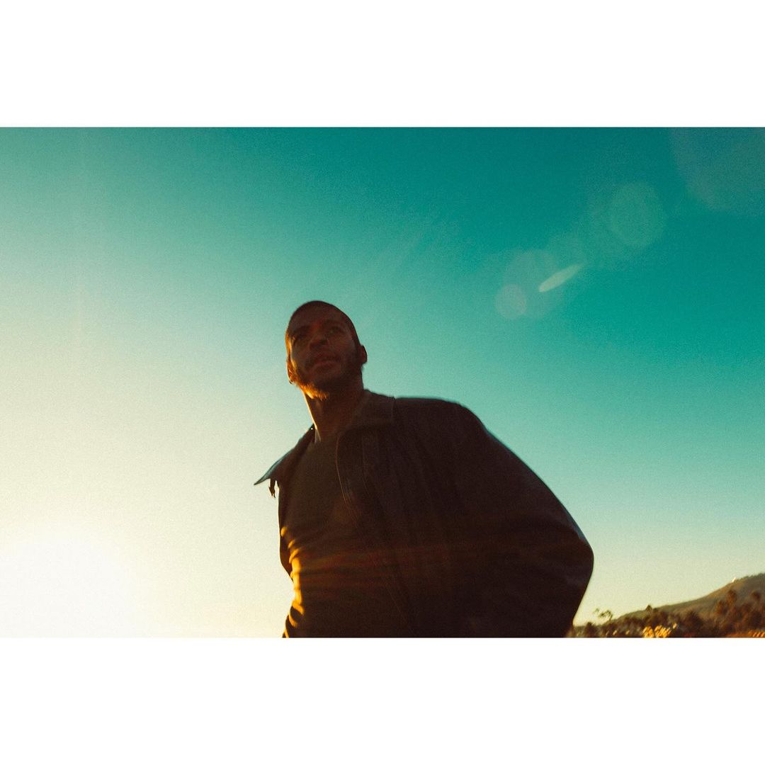 Low-angle shot of an adult male in a jacket against a bright blue sky near a coastal landscape with golden sunlight and a few trees.