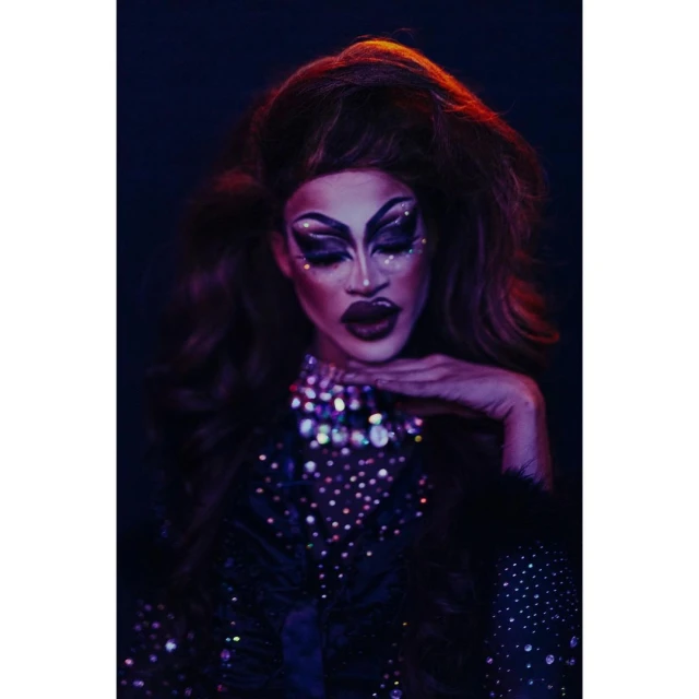 Portrait of drag queen with elaborate makeup, jewel-encrusted collar, and voluminous hair against dark background in an indoor setting.