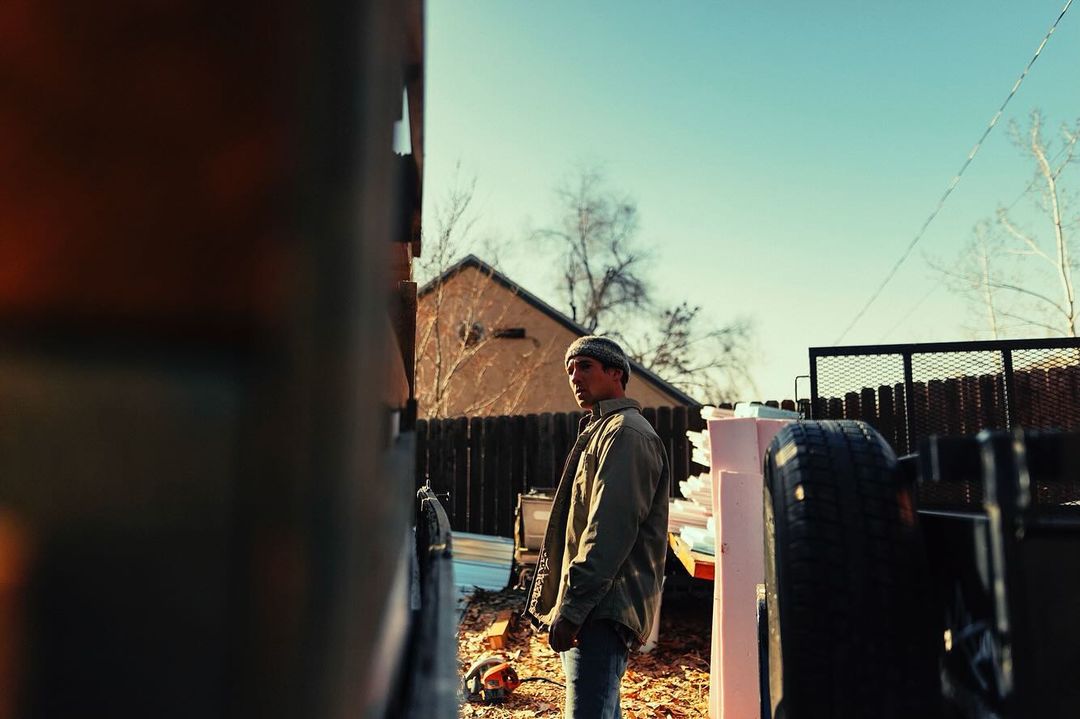 Man in beanie and jacket stands outdoors on construction site in a yard with building supplies under a blue sky on a sunny day.