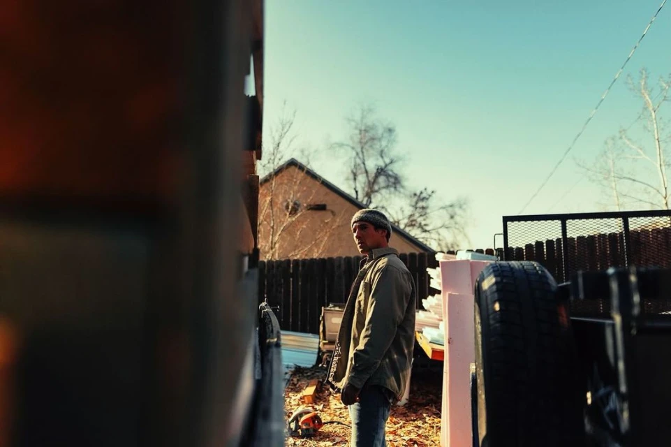 Man in beanie and jacket stands outdoors on construction site in a yard with building supplies under a blue sky on a sunny day.
