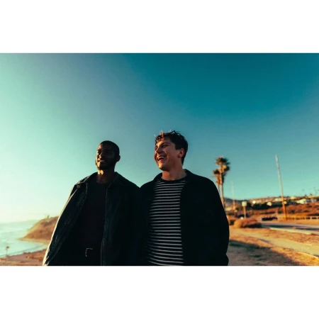 Two men laughing together outdoors against a clear sky backdrop with a coastline visible in the distance.