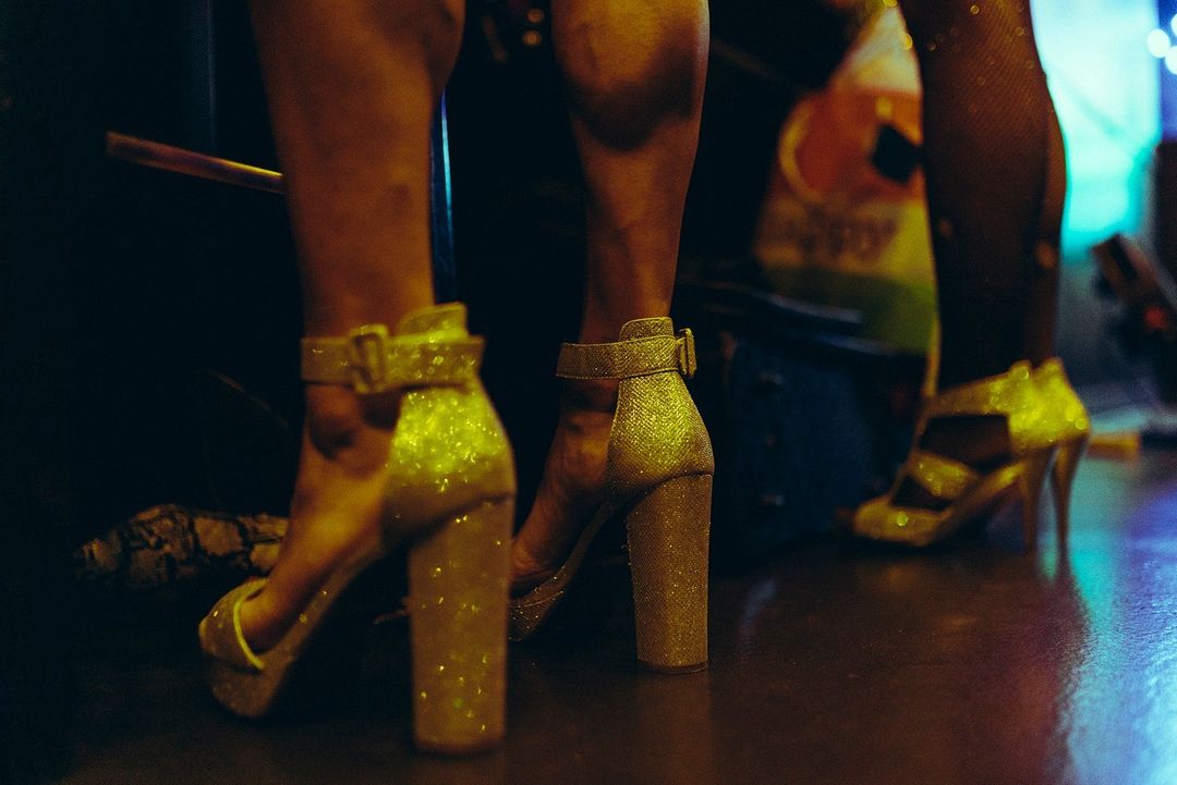 Close-up of two female figures wearing glittering gold high heels with ankle straps, standing indoors under dim, colorful lighting.
