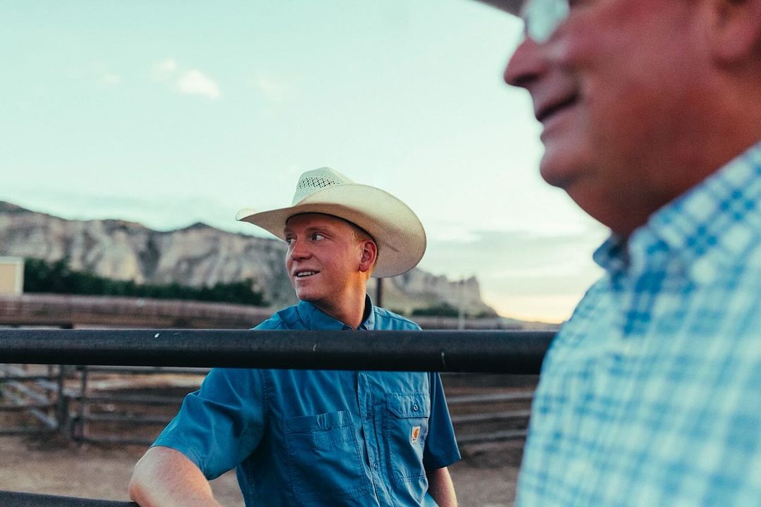 Two men in cowboy hats outdoors. The younger one in a blue shirt is in focus, with mountains visible in the background.