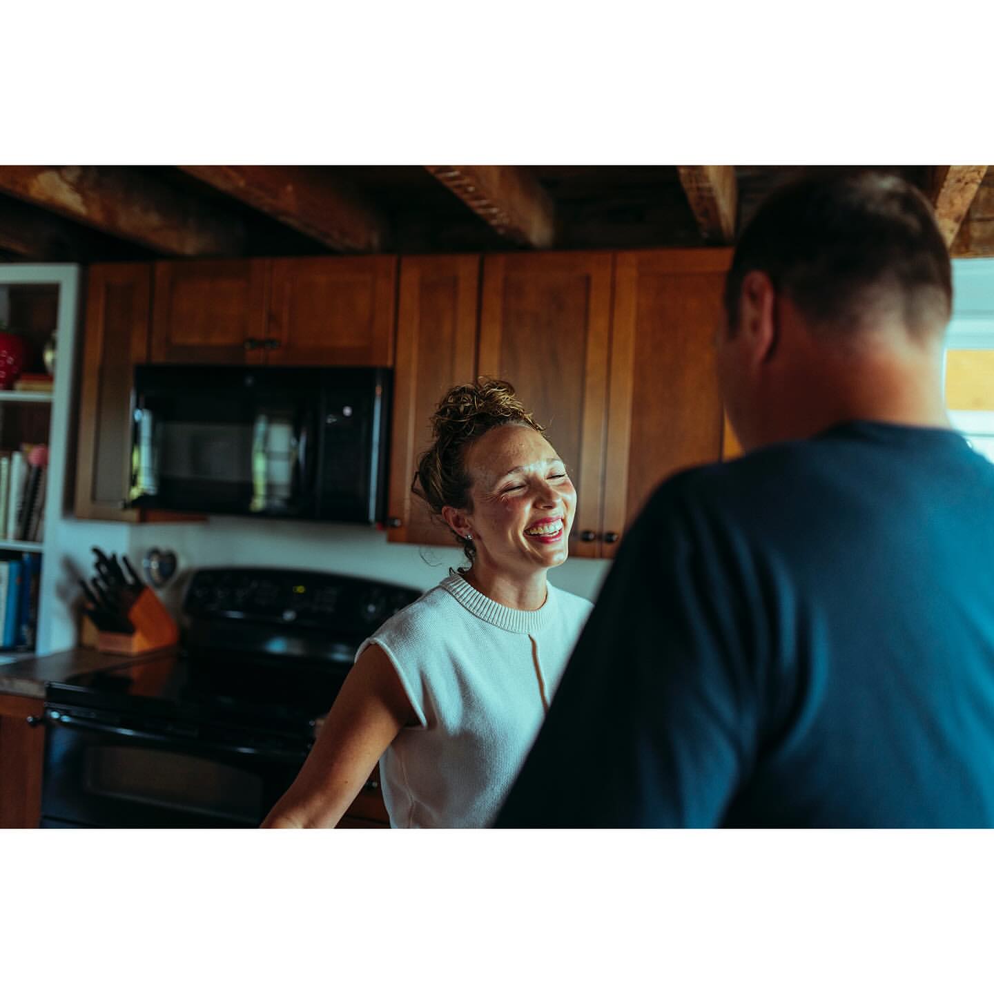 Woman with braces smiles in bright kitchen with wood cabinets while talking to a man. Natural light illuminates the scene.