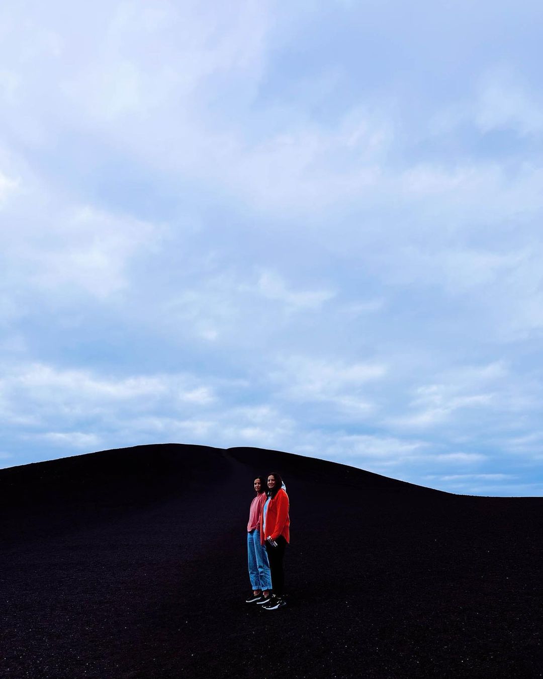 Two people stand in an open landscape of black sand beneath a cloudy sky, dressed in casual clothing with an overcast ambiance.