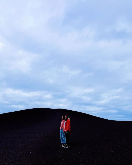 Two people stand in an open landscape of black sand beneath a cloudy sky, dressed in casual clothing with an overcast ambiance.