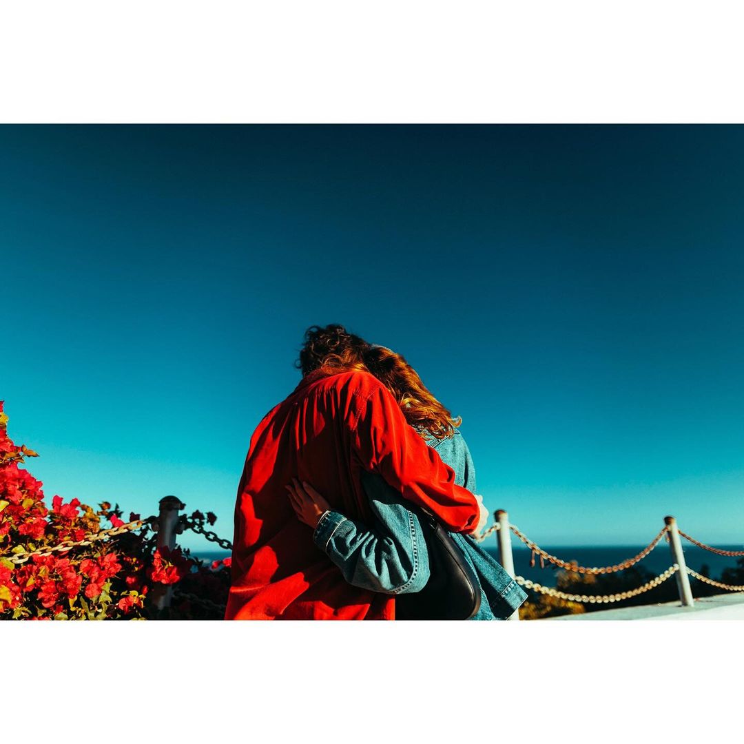 A couple embraces, gazing at the ocean from a flowered overlook under a bright blue sky, creating a romantic and scenic moment.