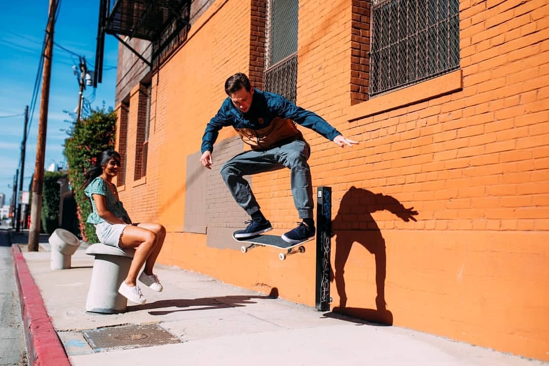 Man performing skateboard trick near orange brick wall while a woman sits nearby. Urban street scene with sidewalk and buildings.